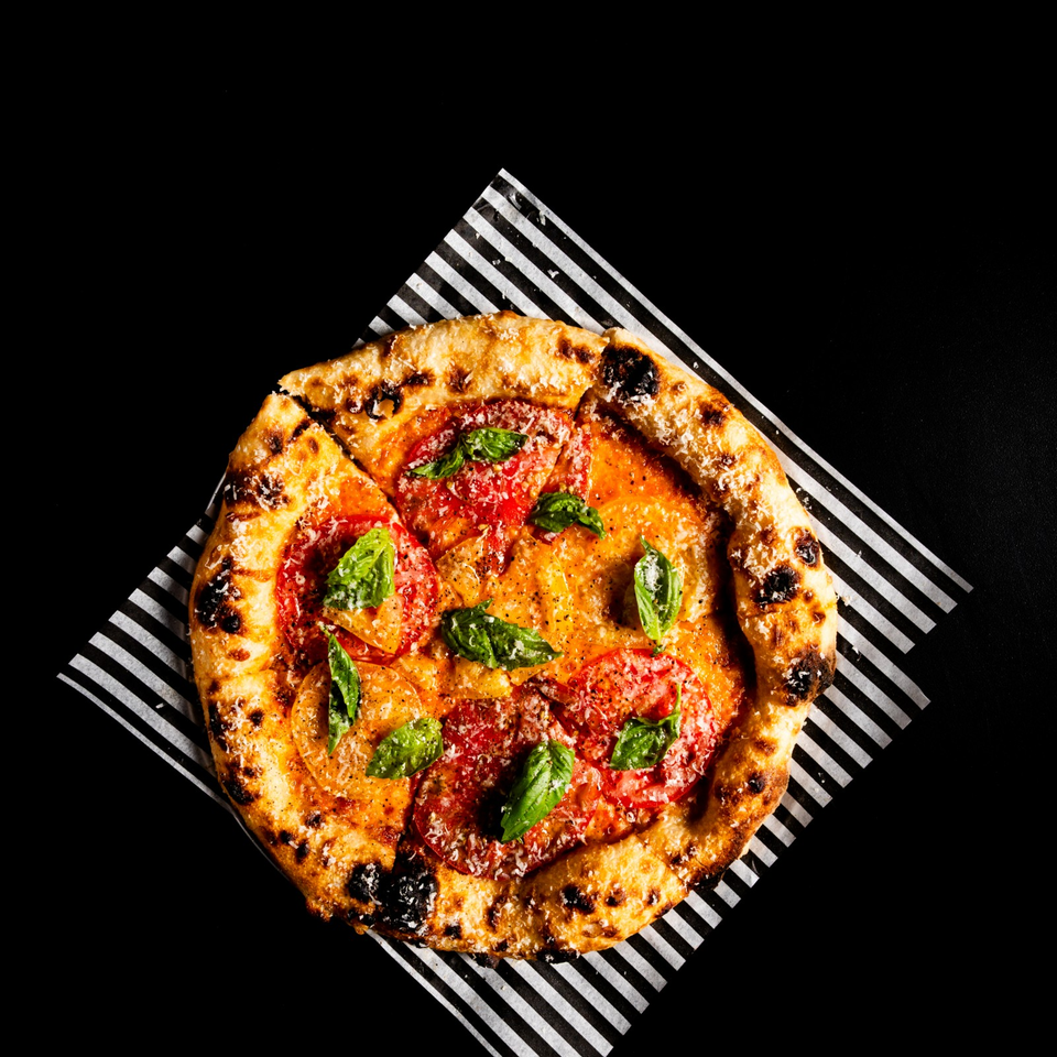 Neapolitan-style pizza topped with tomatoes, basil, grated cheese, and a charred golden crust on striped paper.