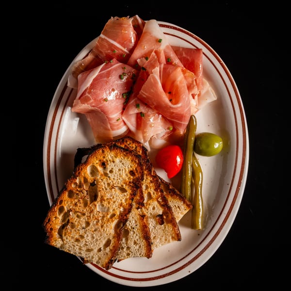 Appetizer plate with prosciutto, grilled sourdough, pickled green beans, red pepper, and an olive.