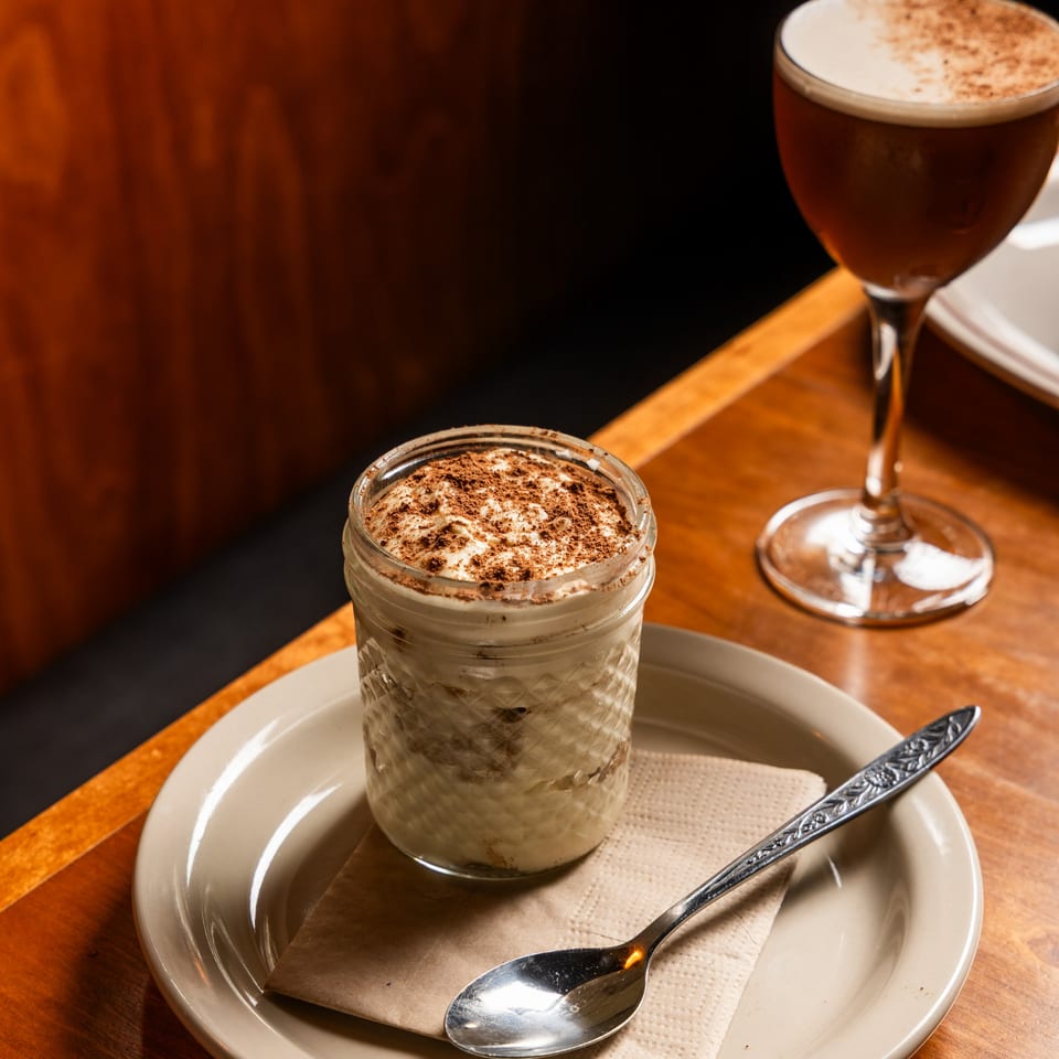 Tiramisu in a glass jar with a spoon, on a plate next to a cocktail on a wooden table.