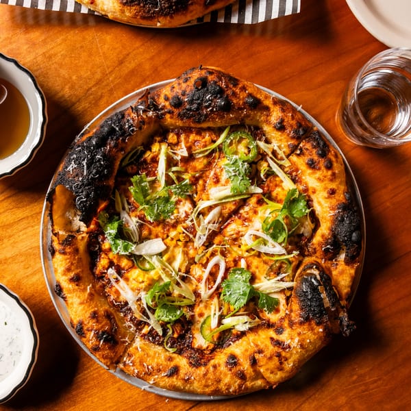 Wood-fired pizza with dark crust, corn, green onions, cilantro, and jalapeños on a wooden table.